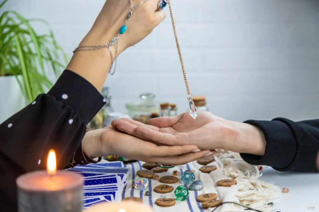 Hands engaging in a pendulum reading over an open palm, surrounded by tarot cards, rune stones, and a lit candle, creating a mystical atmosphere.
