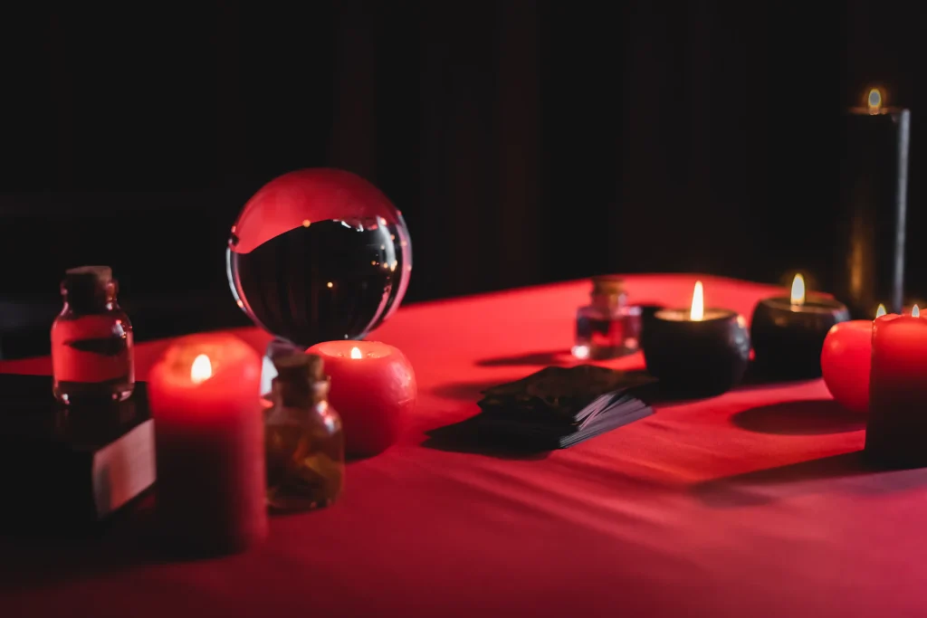 A dimly lit table with a red cloth, featuring a crystal ball, candles, tarot cards, and small potion bottles. The mood is mystical and mysterious.