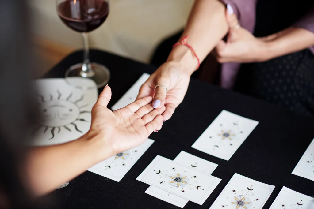 Two hands engaged in a palm reading over a table with tarot cards. A glass of red wine sits nearby, creating a mystical and intimate atmosphere.