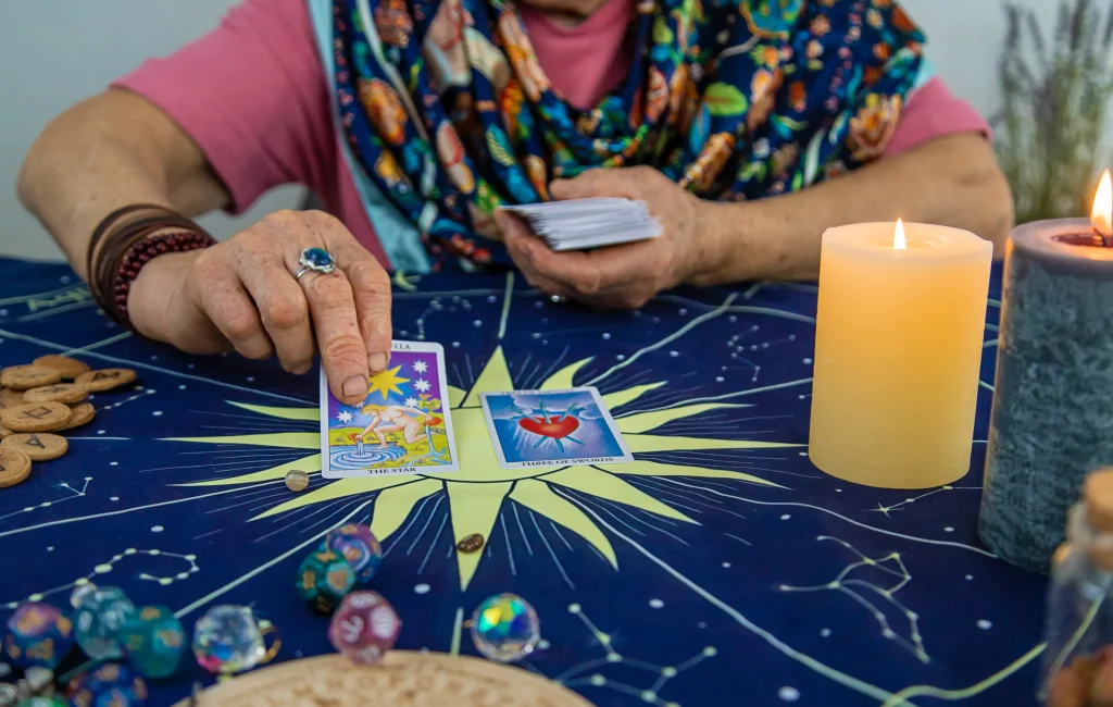 A woman with a colorful scarf lays tarot cards on a starry cloth. The scene includes lit candles and dice, creating a mystical and introspective mood.