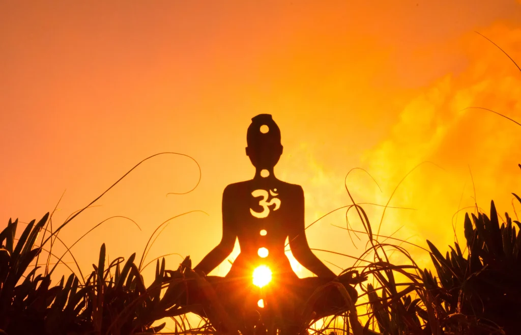 Silhouette of a meditative figure with chakras and an Om symbol at sunset. The warm orange sky creates a serene, spiritual atmosphere.